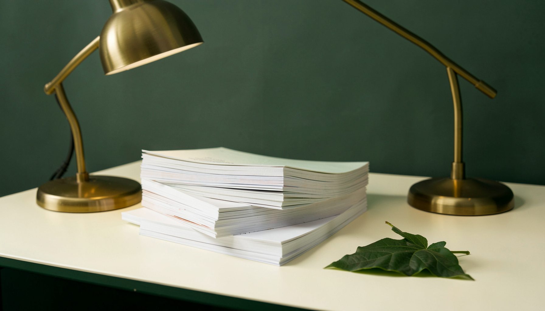 Quiet studio scene of stacked paper booklets, a brass desk lamp, and a pressed leaf on a pale desk in cream and deep green hues