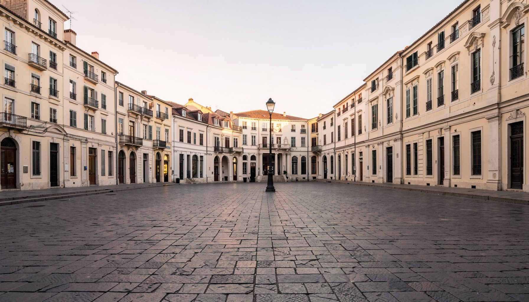 Wide photograph of an empty European plaza at dawn with stone paving, a single lamp post, and soft warm light over pale facades